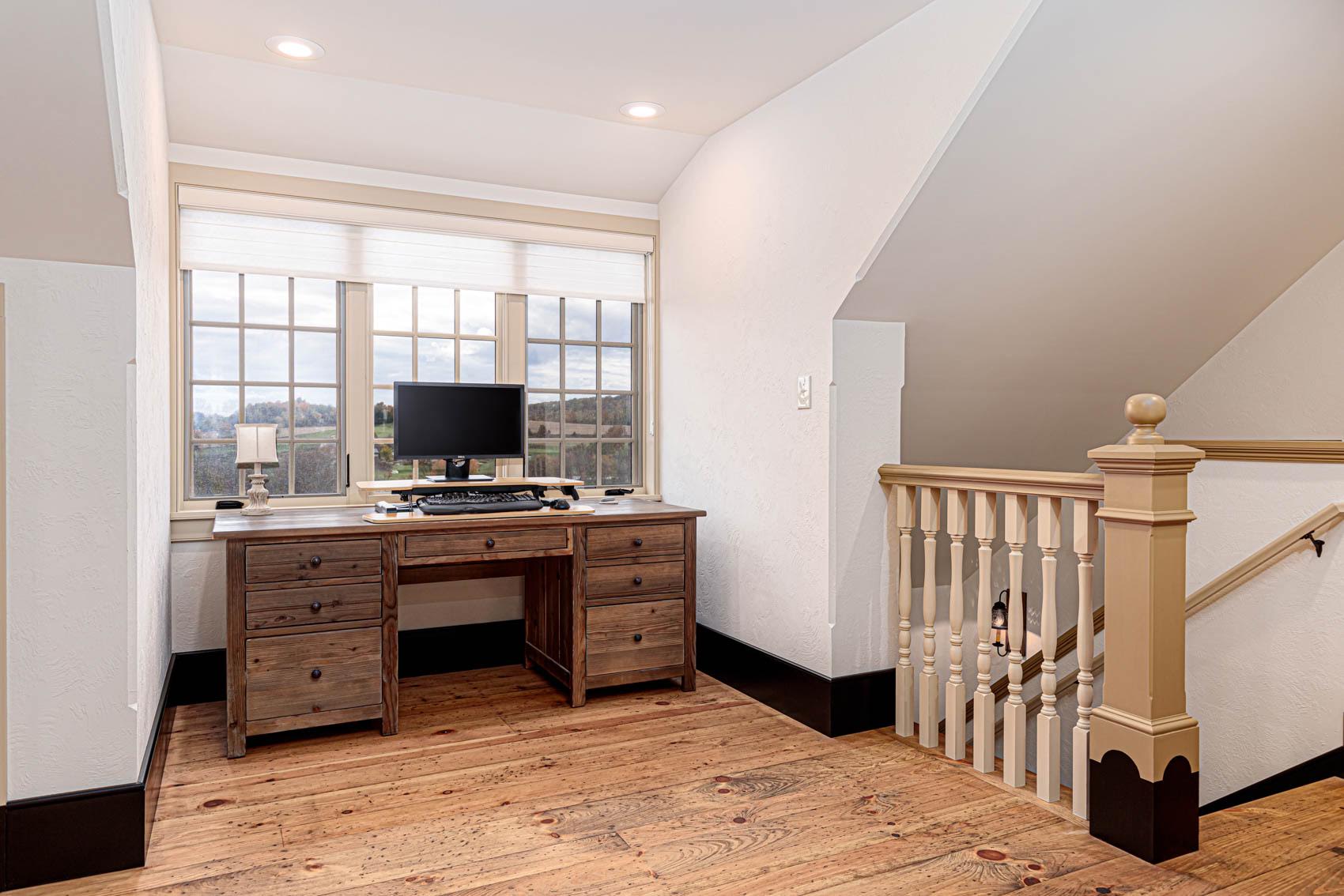 Bright home office space on a landing with a rustic wooden desk, computer setup, large window offering scenic views, and natural wood flooring, next to a staircase with a decorative banister.