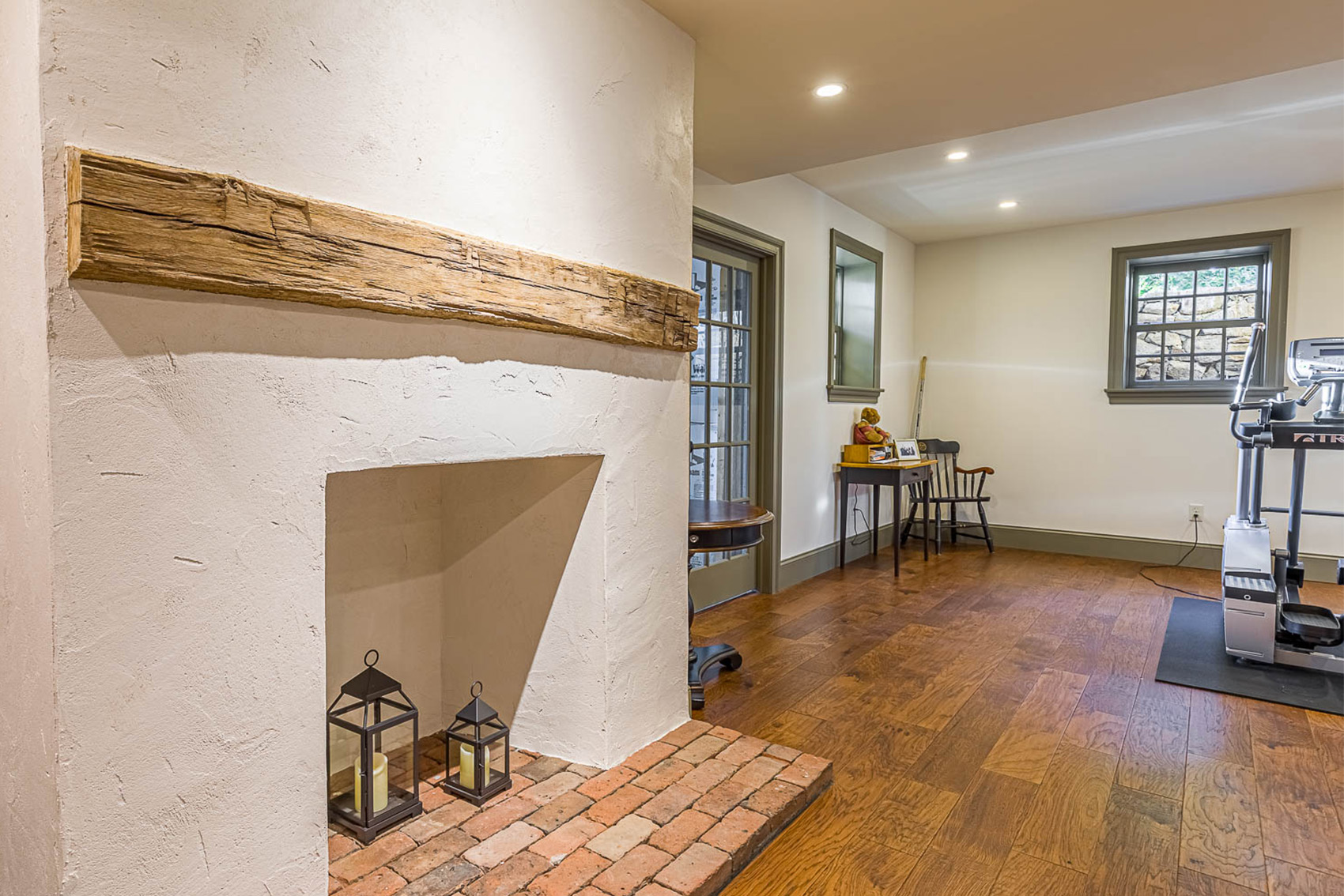Cozy interior room with rustic fireplace featuring a wooden mantel, two lanterns on a brick hearth, hardwood flooring, and a small fitness area with exercise equipment