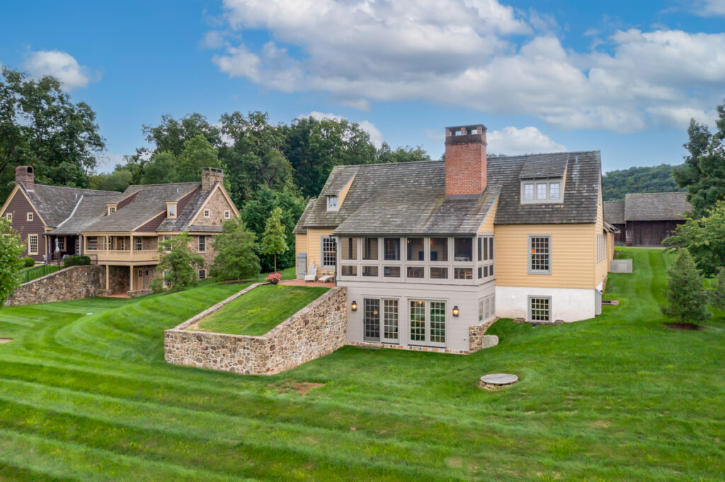 Restored historic home with stone foundation, yellow wood siding, and large chimney, set on a lush green lawn with mature trees and a scenic countryside backdrop.
