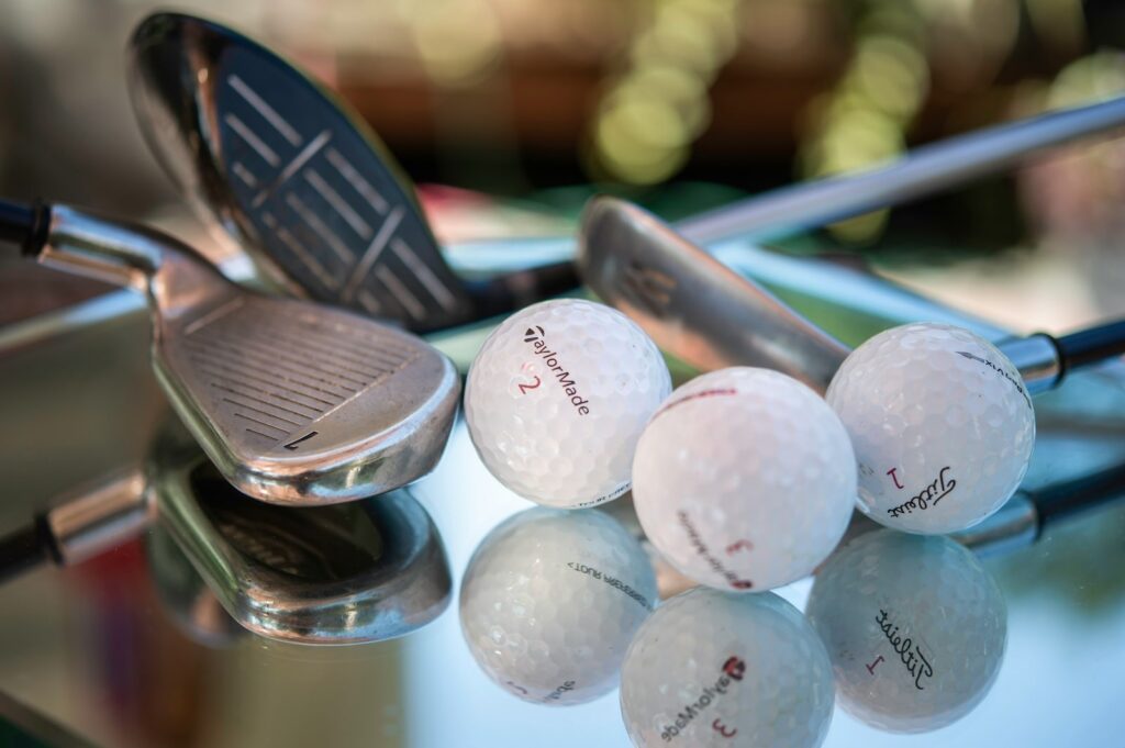 Close-up of golf clubs and white golf balls, including TaylorMade and Titleist brands, resting on a reflective glass surface with a blurred background.