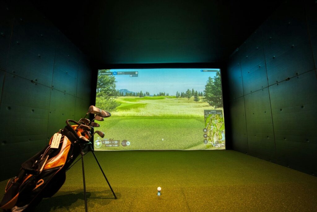 Indoor golf simulator setup with a virtual fairway projected on a screen, a golf bag with clubs on the left, and a tee and golf ball positioned in front.