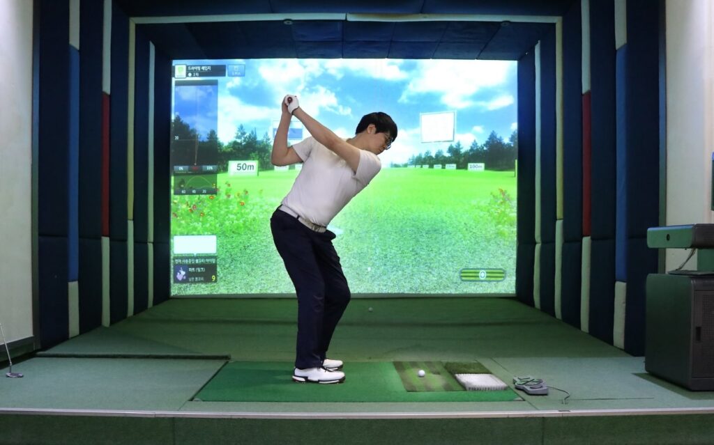 Man practicing golf indoors using a golf simulator, standing in mid-swing in front of a large virtual screen displaying a golf course.